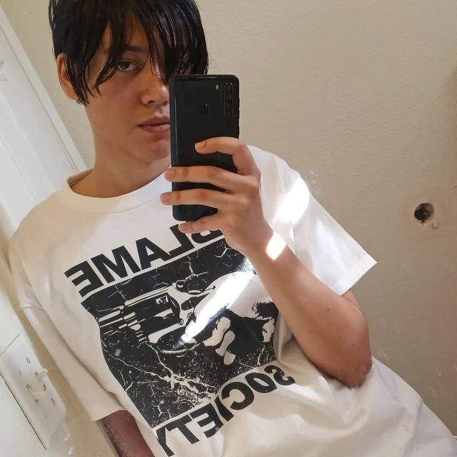 Person wearing a white t-shirt with black graphics and text, holding a phone.